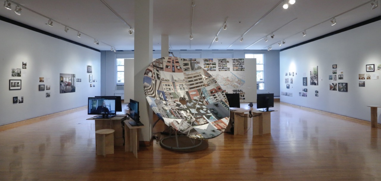 A full gallery with photographs arranged loosely on the walls and a large satellite shaped structure in the center flanked by television monitors on wooden stands