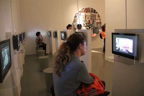people sit on pedestals and watch individual television monitors playing videos in a gallery
