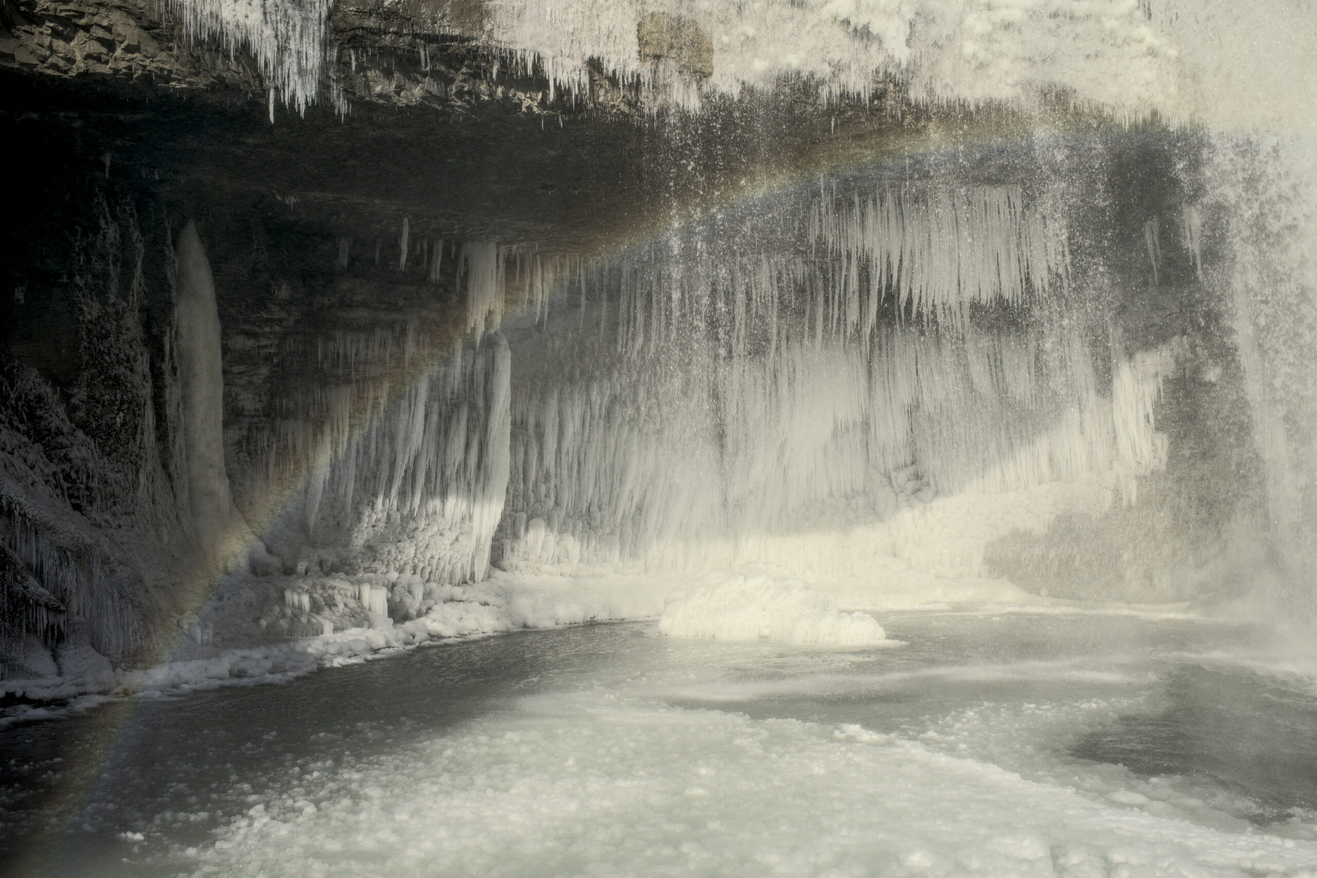A frozen waterfall in the background is illuminated by the sun to create a faint rainbow with snow in the foreground