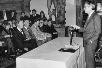A man speaking at a table and microphone to seated audience.