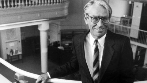 A white-haired man wearing eyeglasses stands on a balustrade.