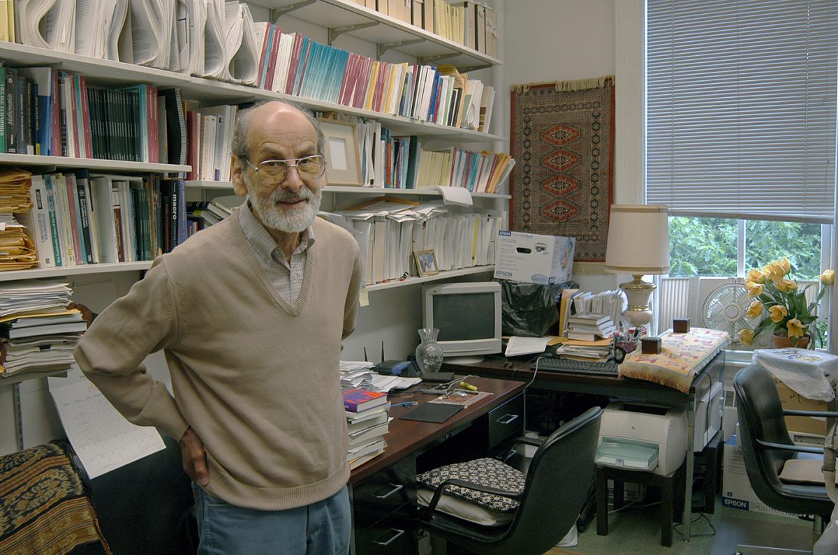 A bearded man wearing glasses stands near bookshelves and a computer
