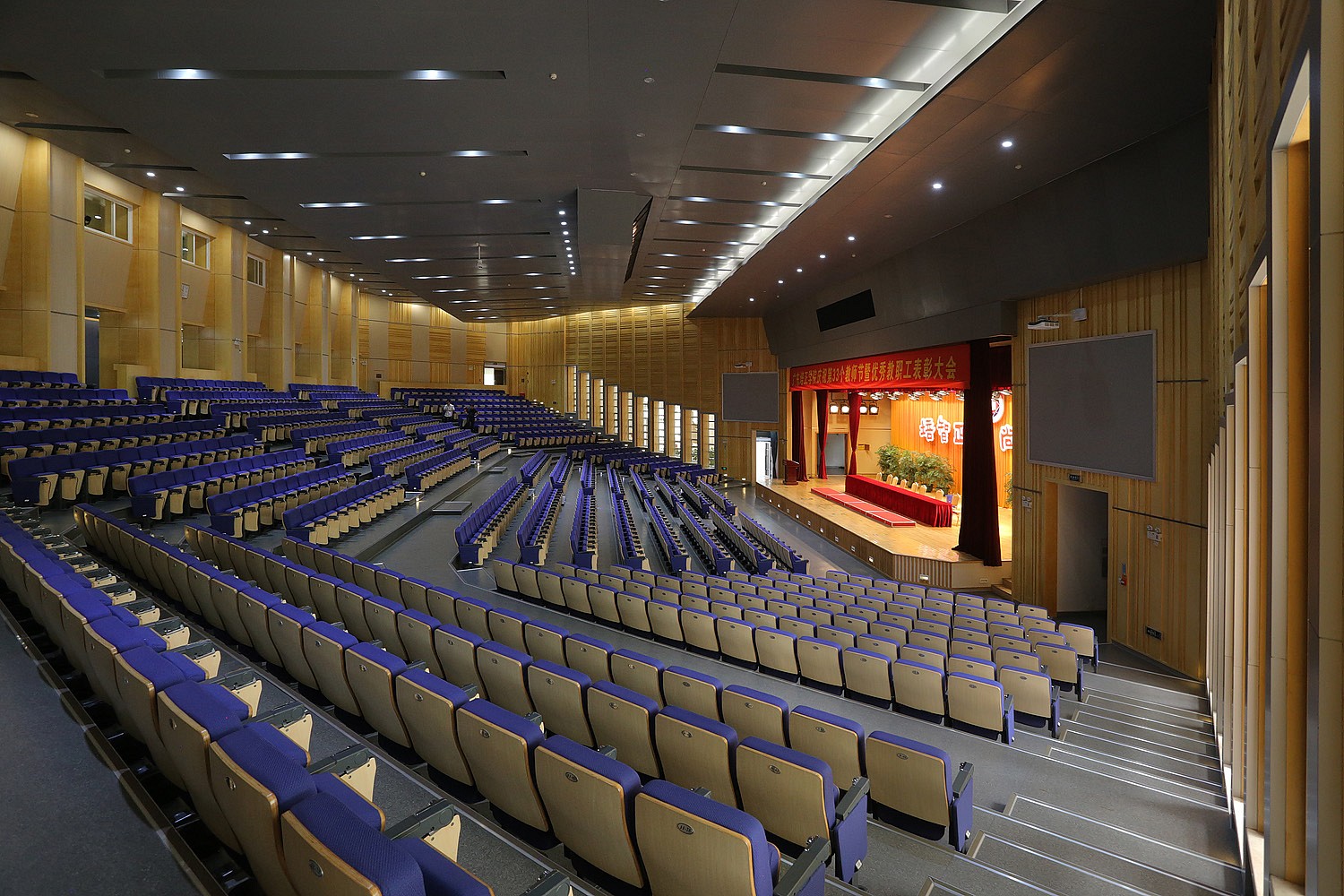 Blue seats in a large, empty auditorium face a lit stage.
