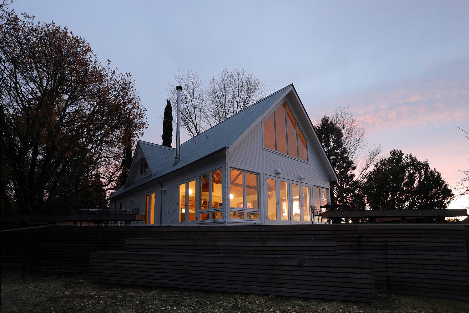 An exterior view of a white house with warm indoor lighting seen through the many glass doors and windows in the evening.