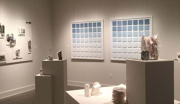 White toned installation with various small sculpture pieces scattered on pedestals with light blue and gray prints on the walls.