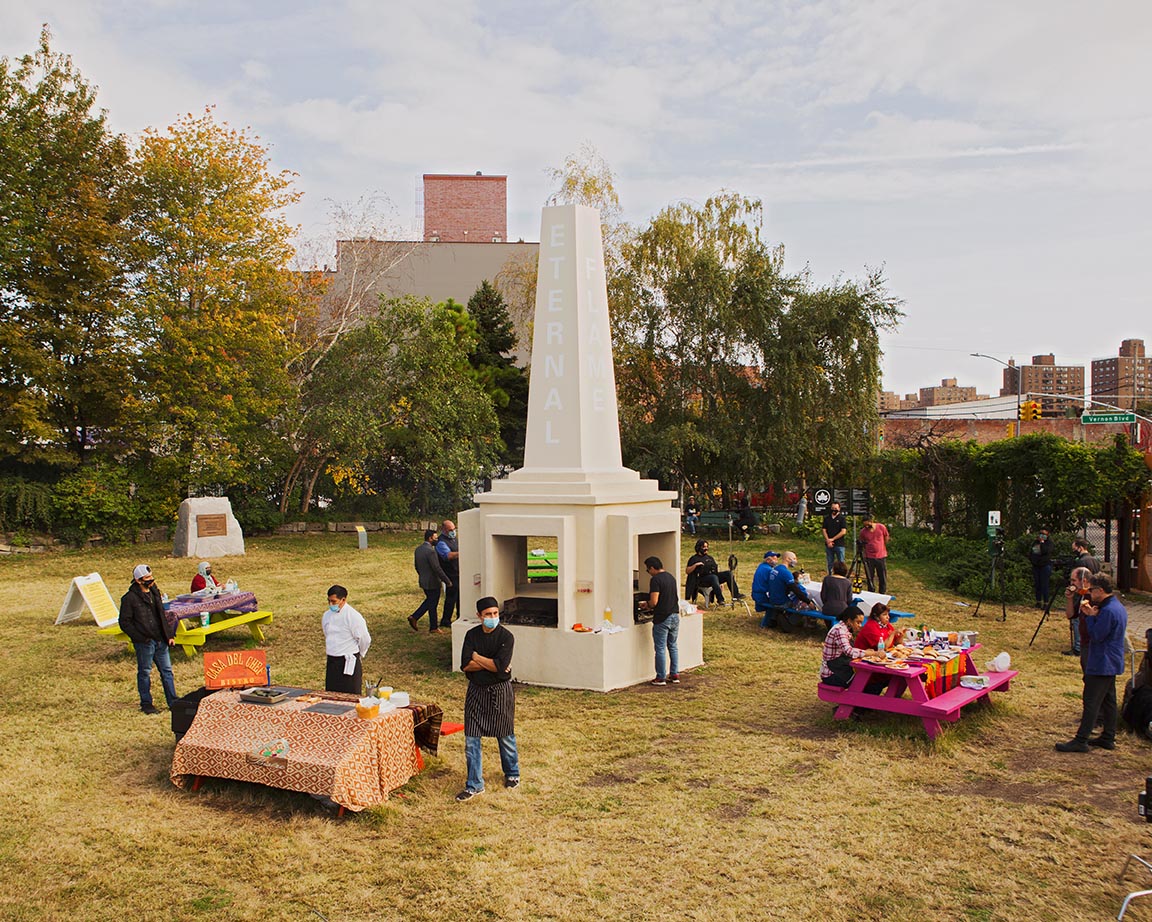A center monument in a park is surrounded by picnic tables in bright colors with people standing at each table.