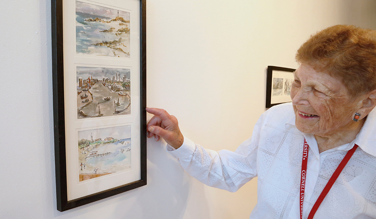 A person with a white shirt and red cornell university lanyard pointing at a frame willed with watercolor paintings.