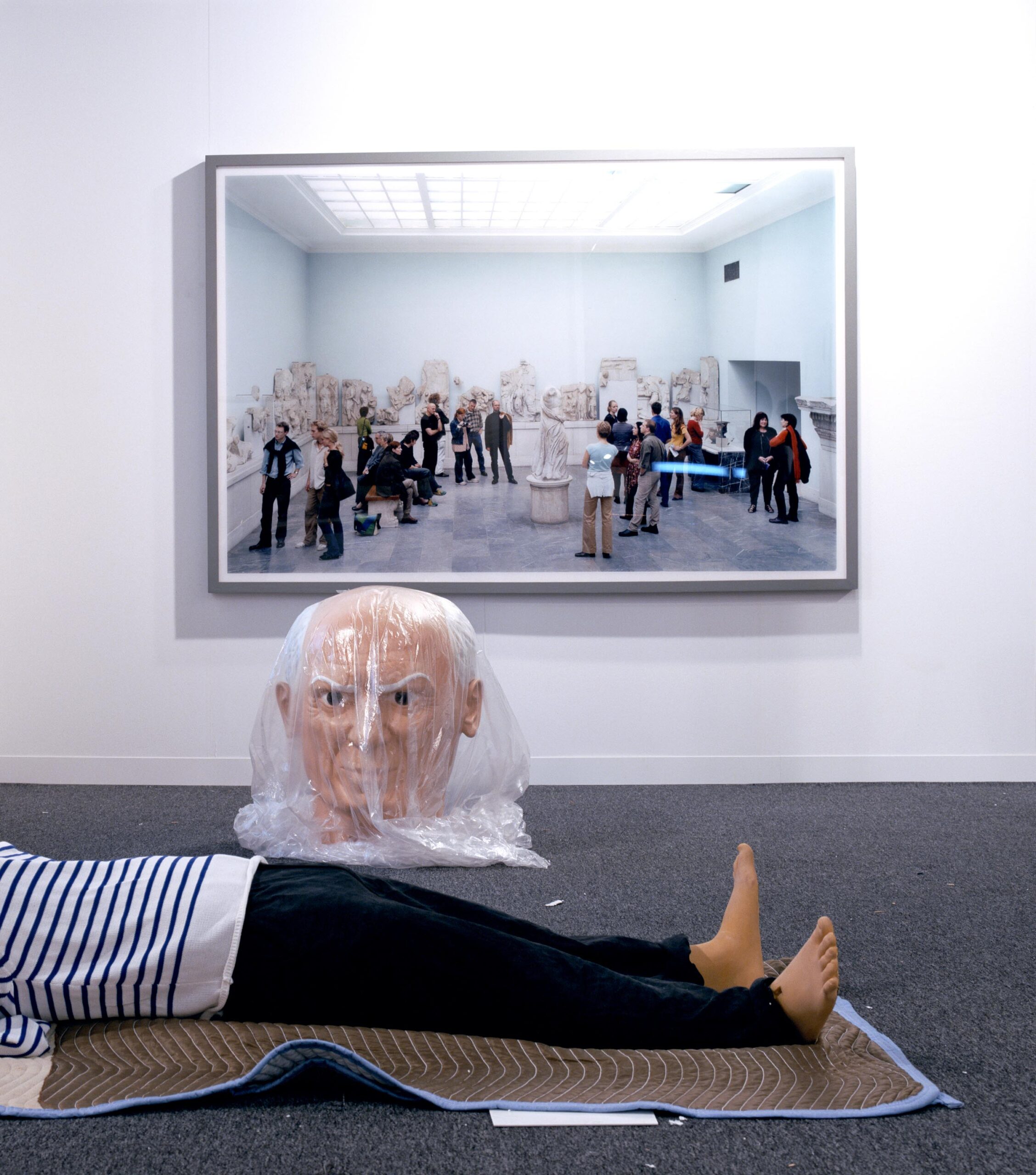 image of a Maurizio Cattelan piece of Picasso figure wrapped, the head is wrapped in plastic and the body is laid on a moving blanket with a photograph in the background