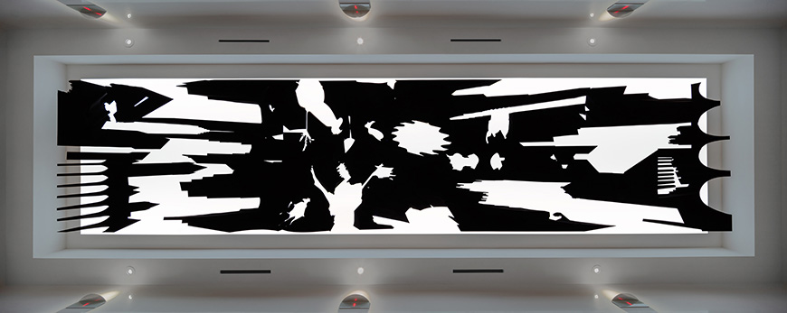 A black and white abstract painting on a large wall in a gallery setting. The painting features bold, angular shapes and lines, creating a dynamic and energetic composition.