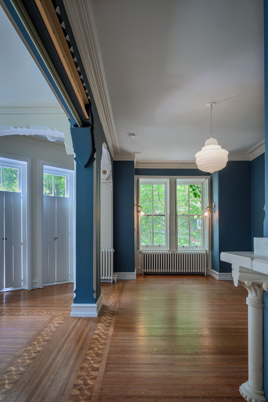 A photo of a spacious, historic room with blue walls, white trim, and a hardwood floor with an intricate inlaid pattern. Large windows with white shutters let in natural light. A white pendant lamp hangs from the ceiling, and there is a radiator against the wall.