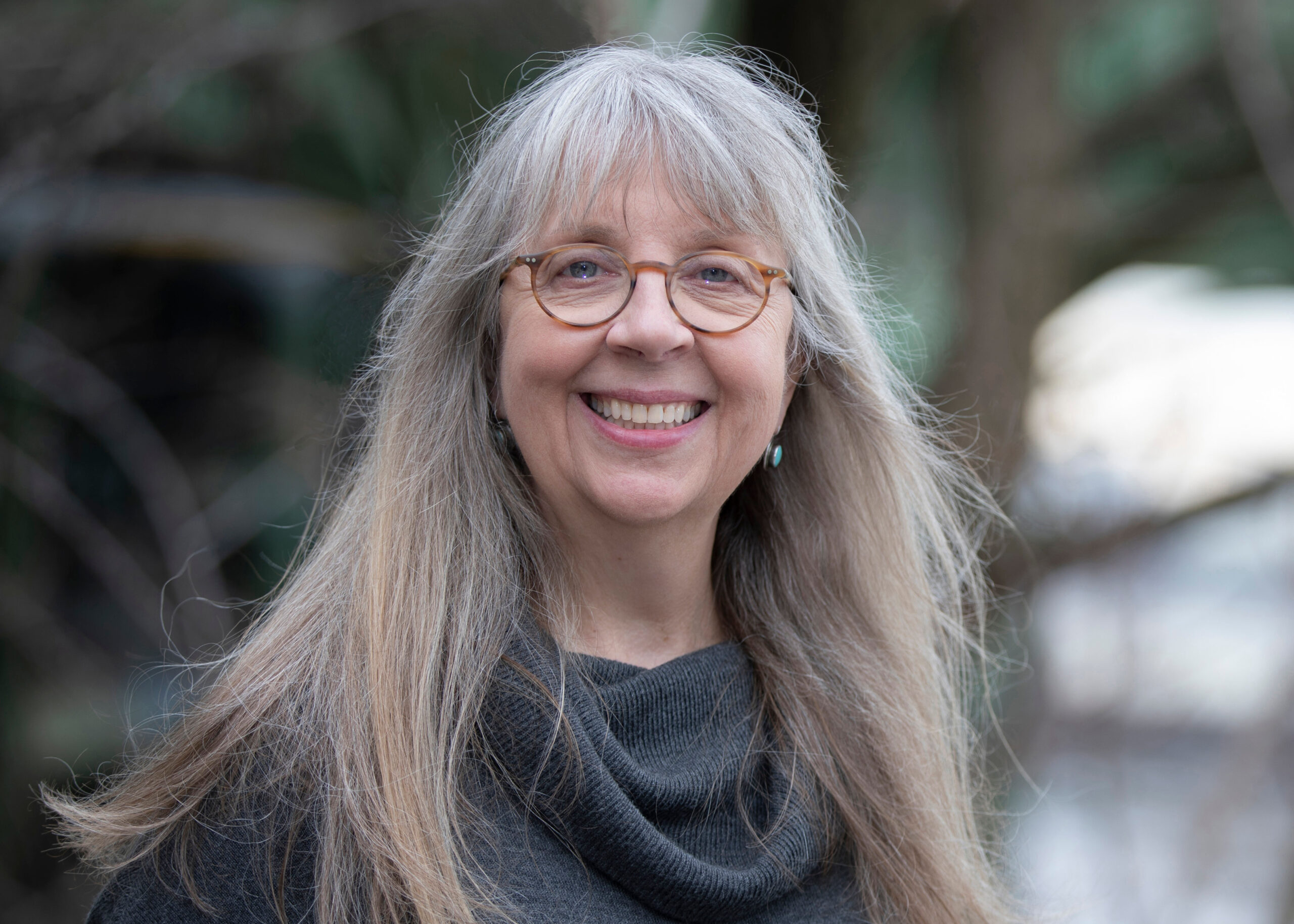 An older woman with long gray hair, wearing glasses with round tortoise shell frames, smiles at the camera.