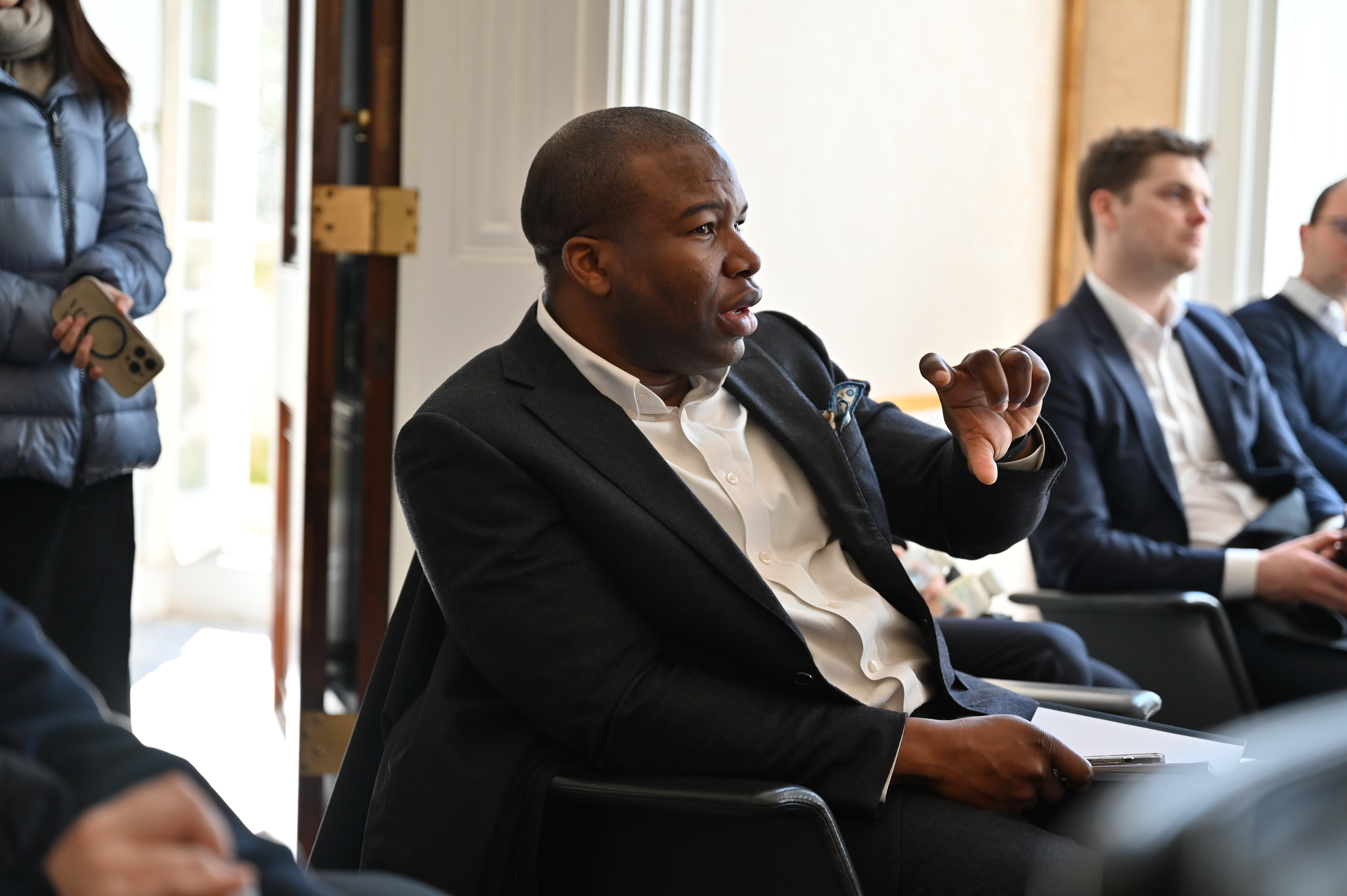 a seated man in a jacket speaking with hand gesture