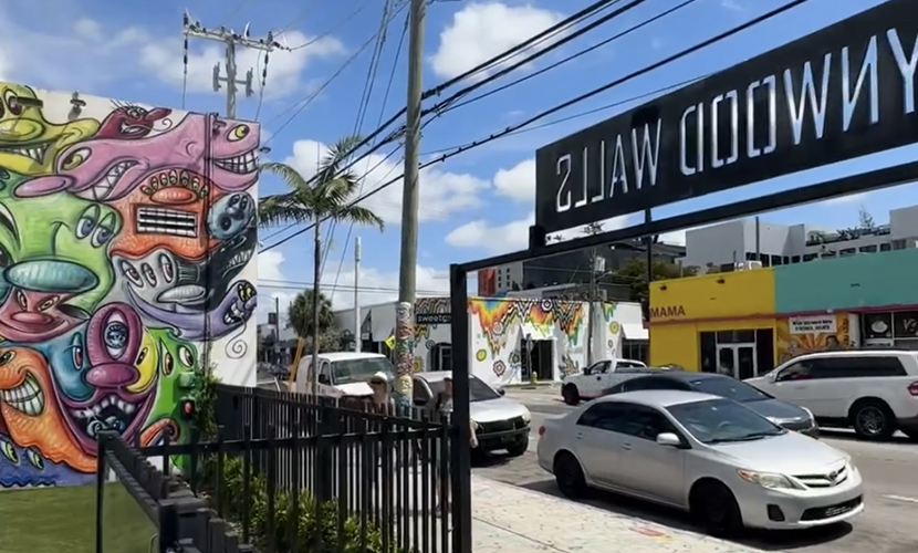 A street scene with a mural, cars, and a Wynwood Walls entry sign