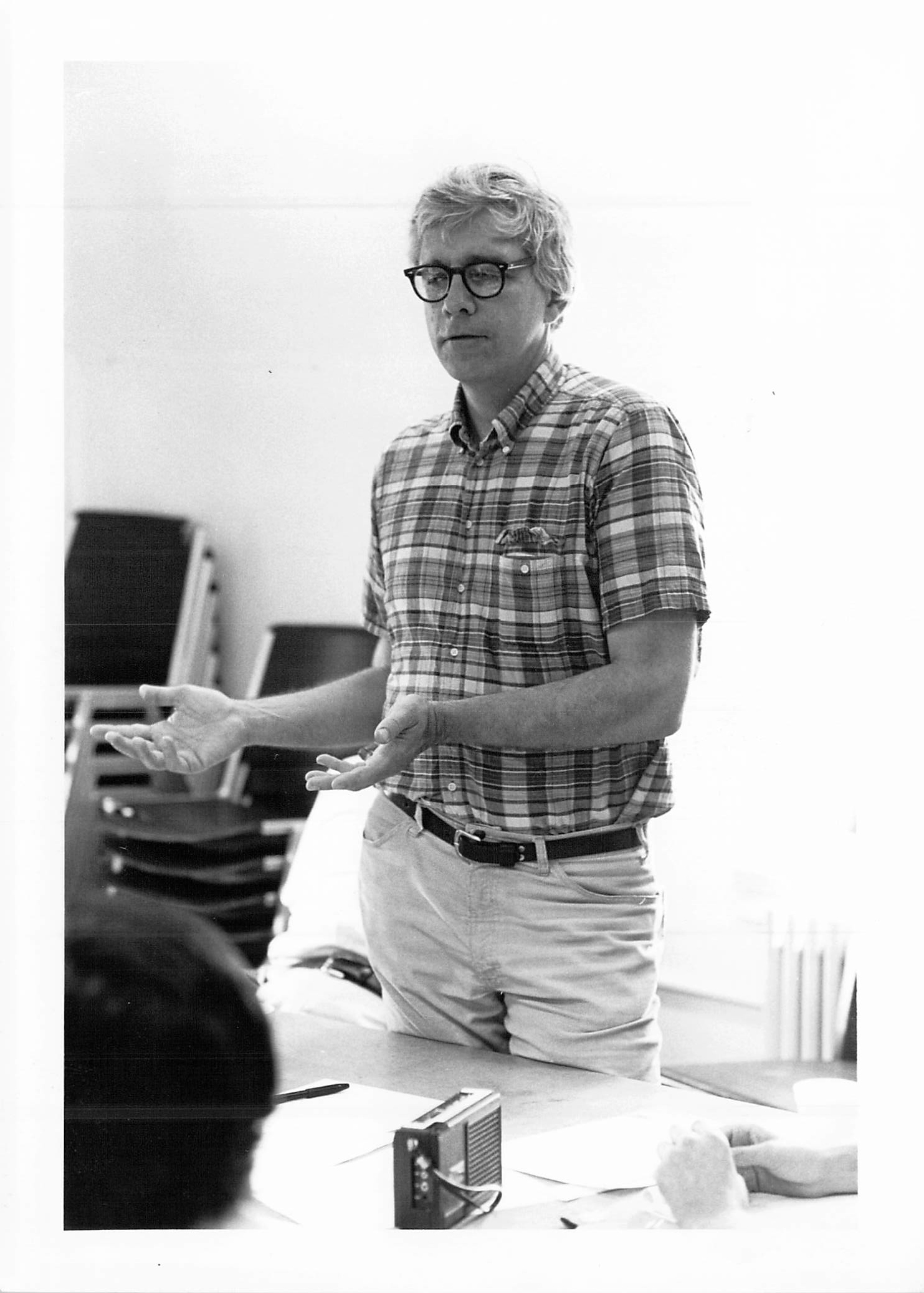 Black and white image of a man gesturing with his hands while speaking in front of a table.