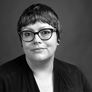 Person with short, dark hair wearing black glasses and a black blouse.