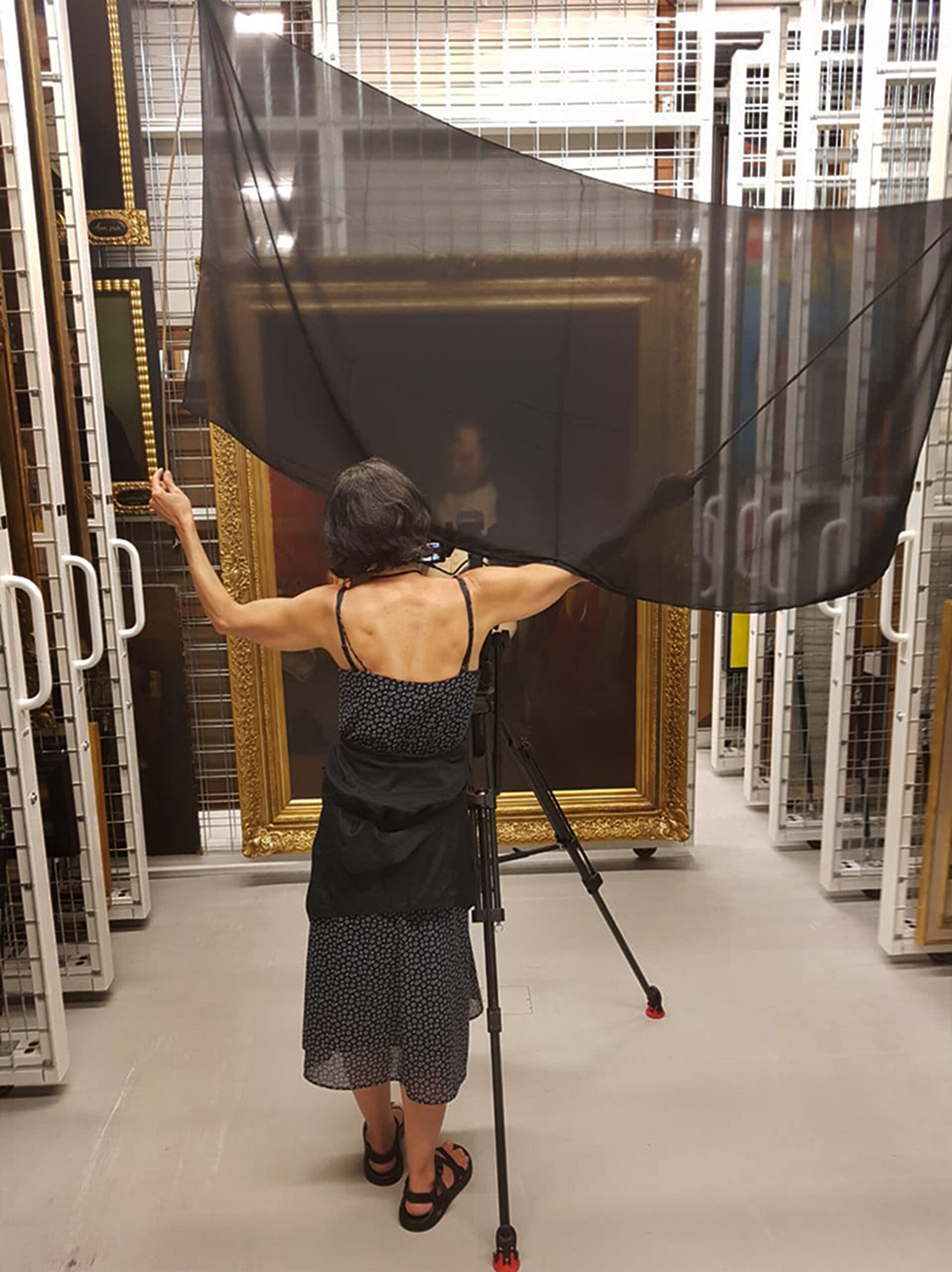 The back of a woman in a black dress faces the camera as she holds a sheet of translucent black fabric in the air. A video camera sits on a tripod, pointing toward a large painting.
