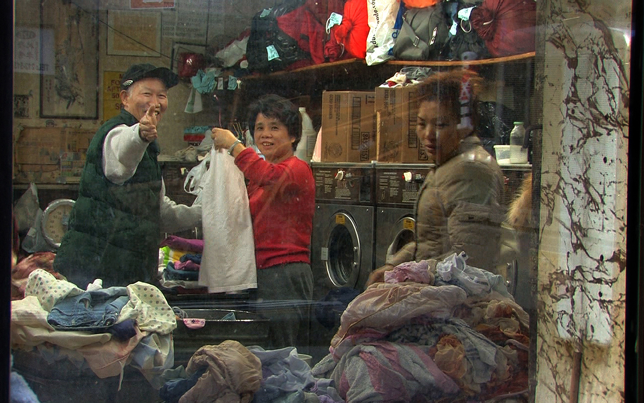 A Chinese women and a Chinese men are folding clothes inside a laundry mat, accompanied by another Chinese woman.