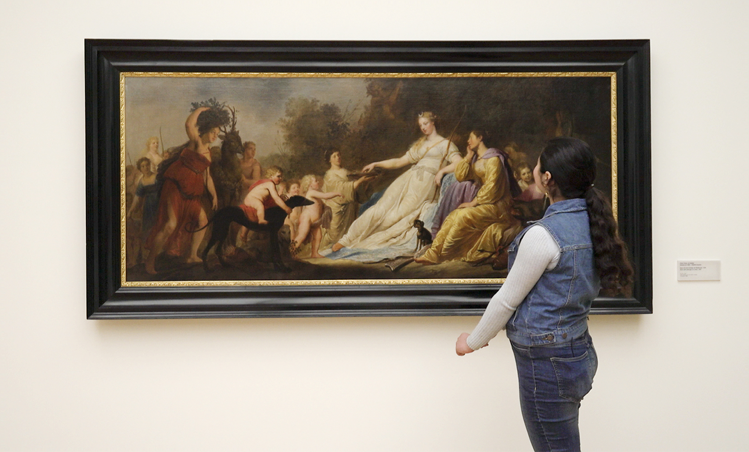 A teenage girl looks at a painting of a woman who is surrounded by adults and being approached by two cherubic children.