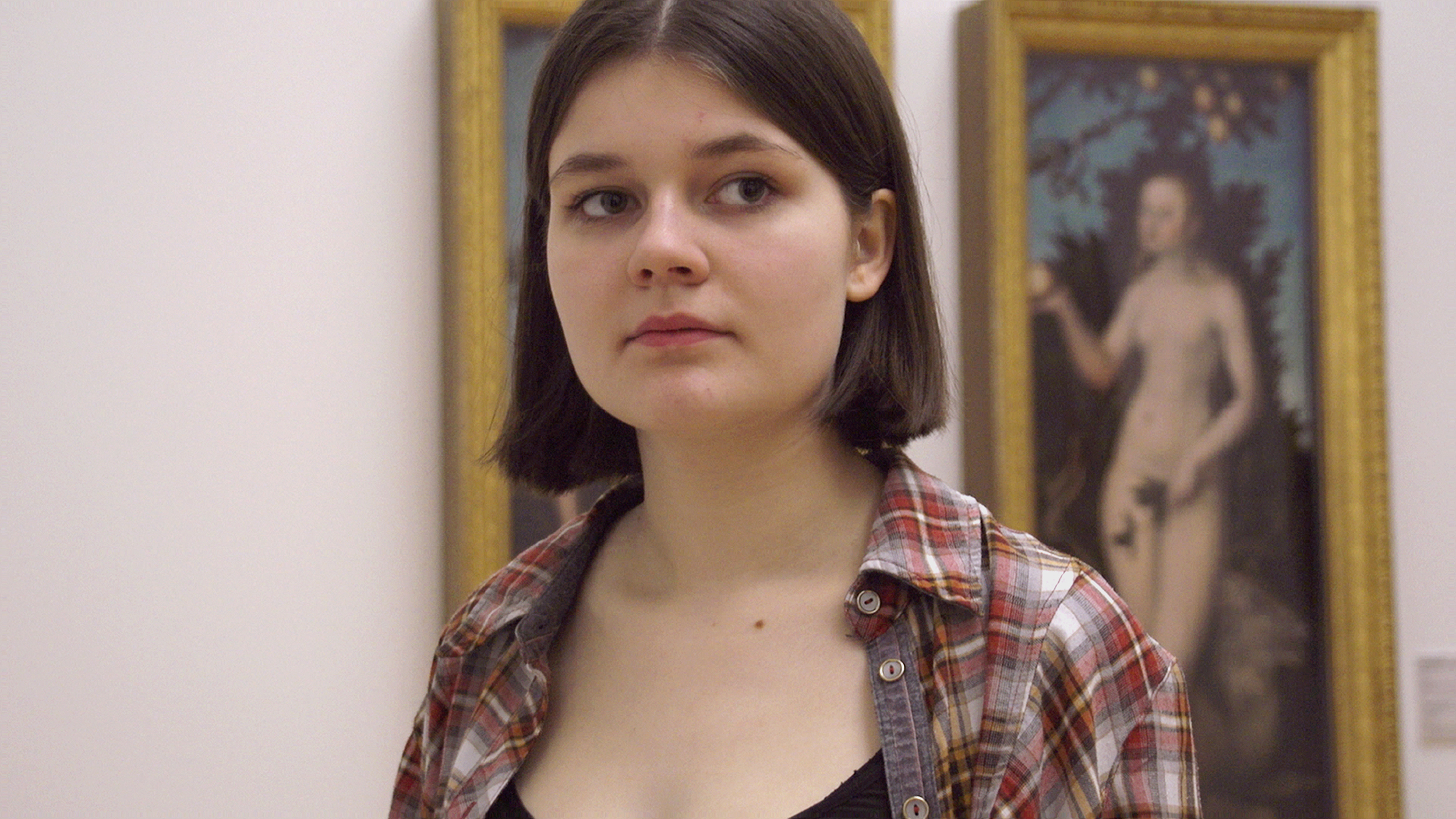 A teen girl gazes off camera with a serious expression while standing in front of a painting of a nude 