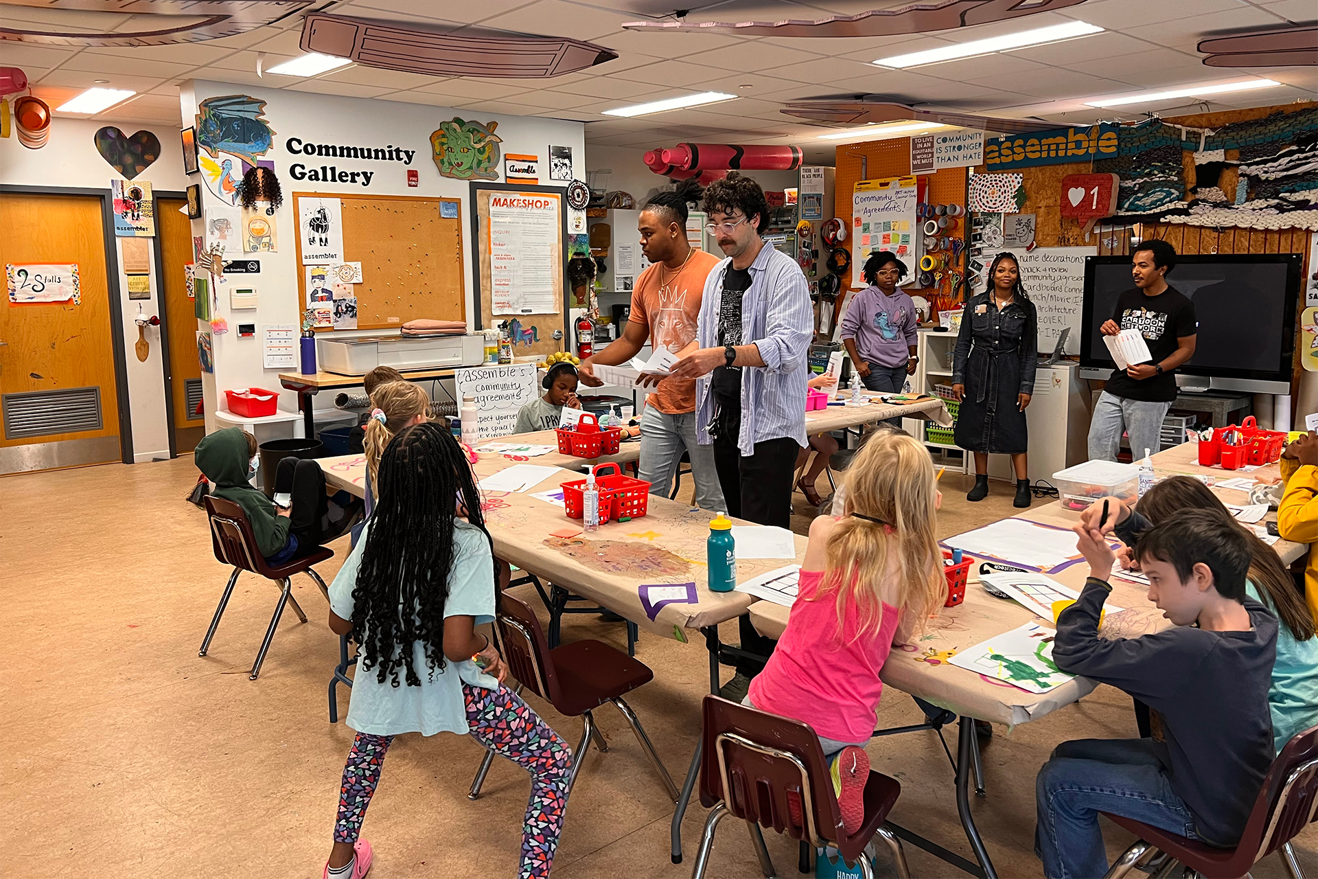 A classroom of elementary students are gathered around tables, being instructed by young adults