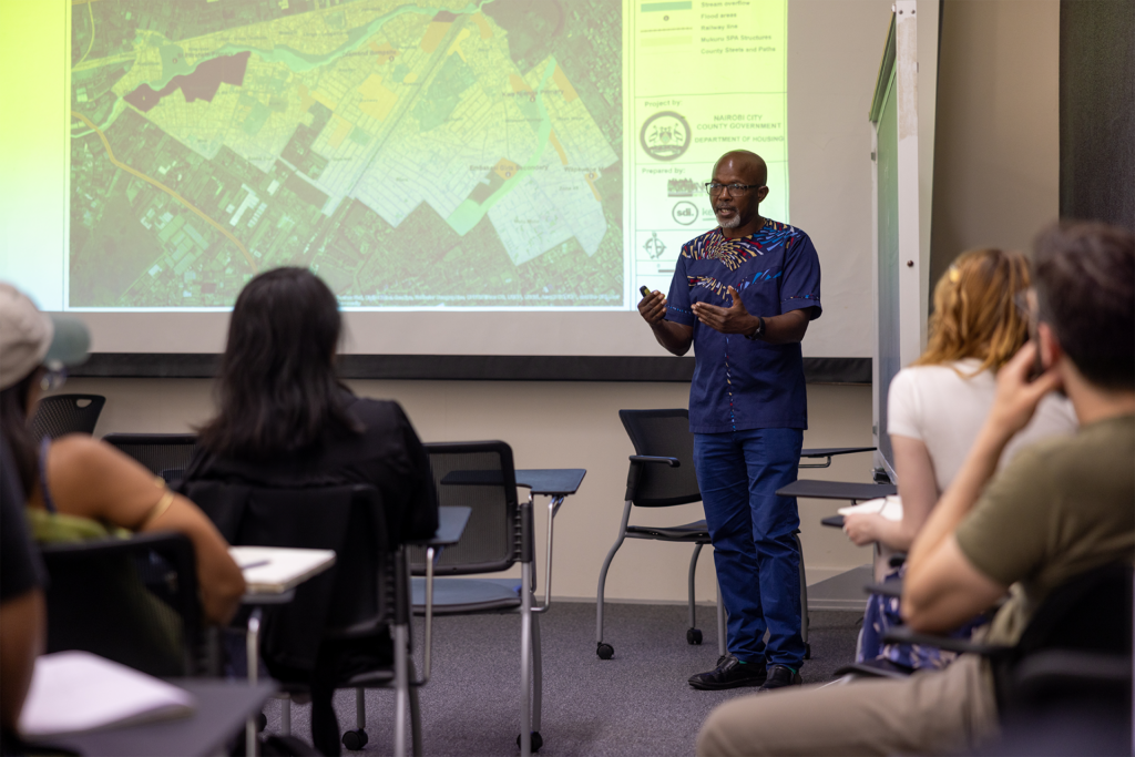 Standing in front of a map projected on a screen, a man addresses a classroom of students