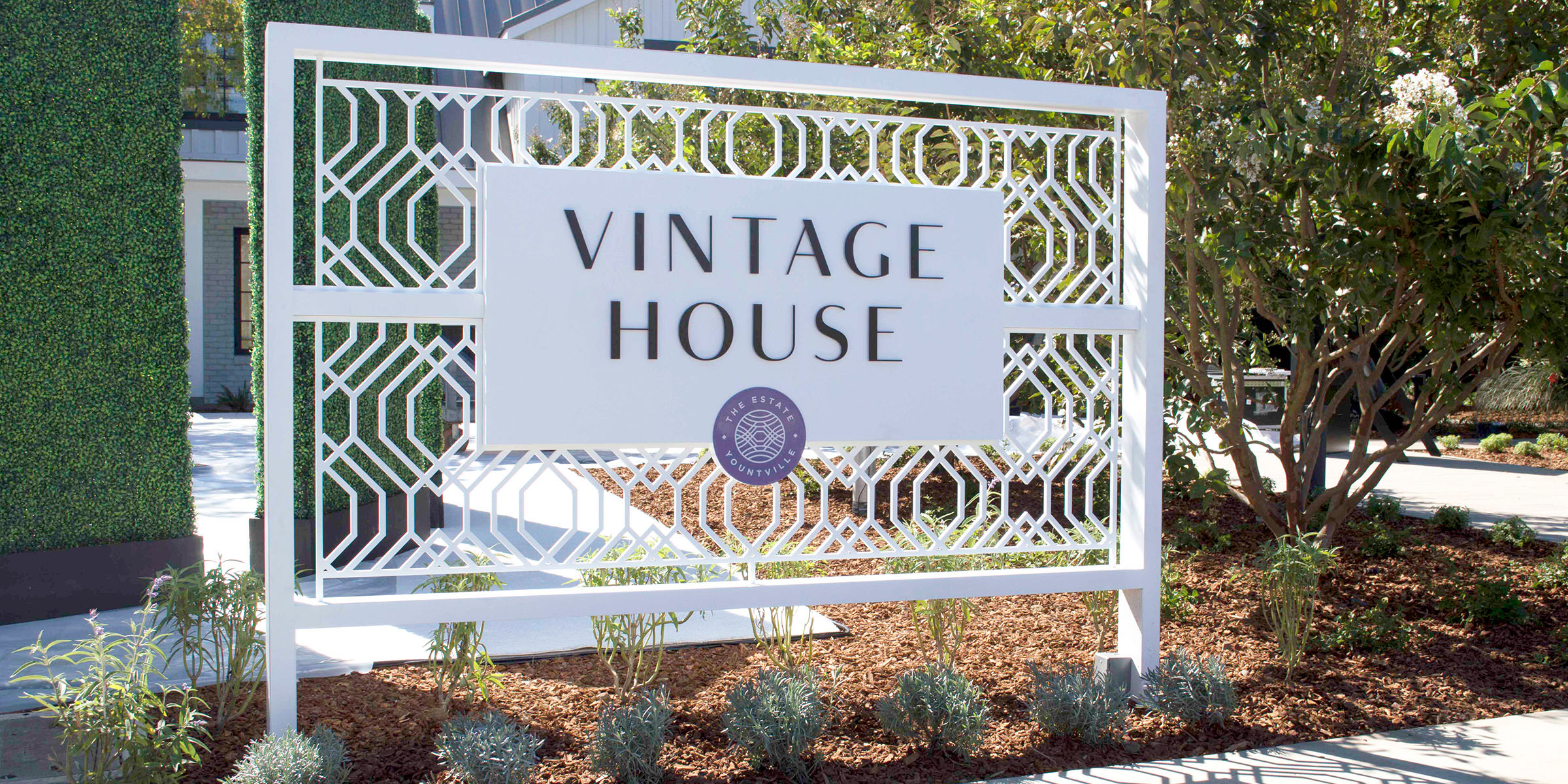 An outdoor white sign is adorned with the words Vintage House in black and a round purple logo for The Estate Yountville