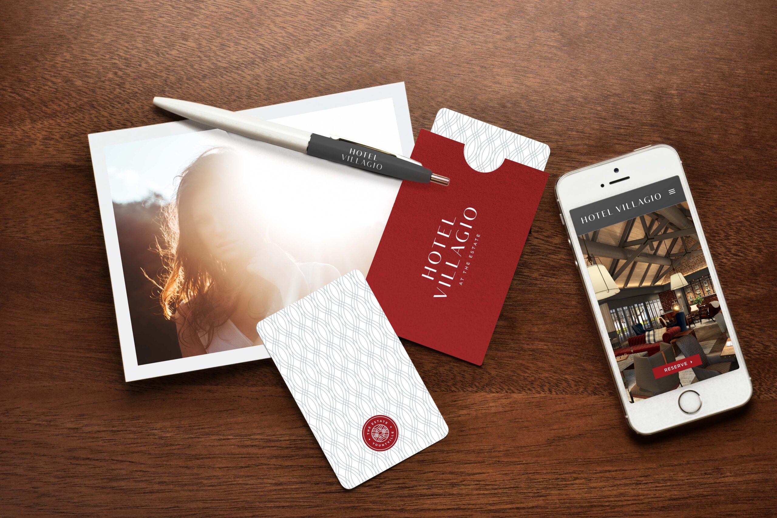 A photo of a woman, a pen, two hotel keycards and a red envelope printed with the words 'Hotel Villagio', and a smartphone are arranged on a wooden desk