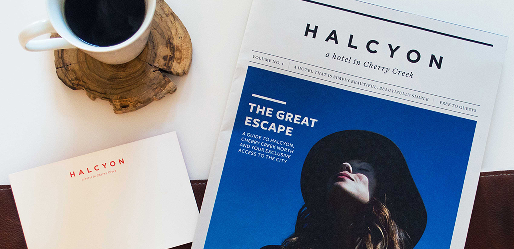 A white mug filled with coffee sits on a wooden coaster on a white table, next to a notepad and a magazine for the Halcyon hotel