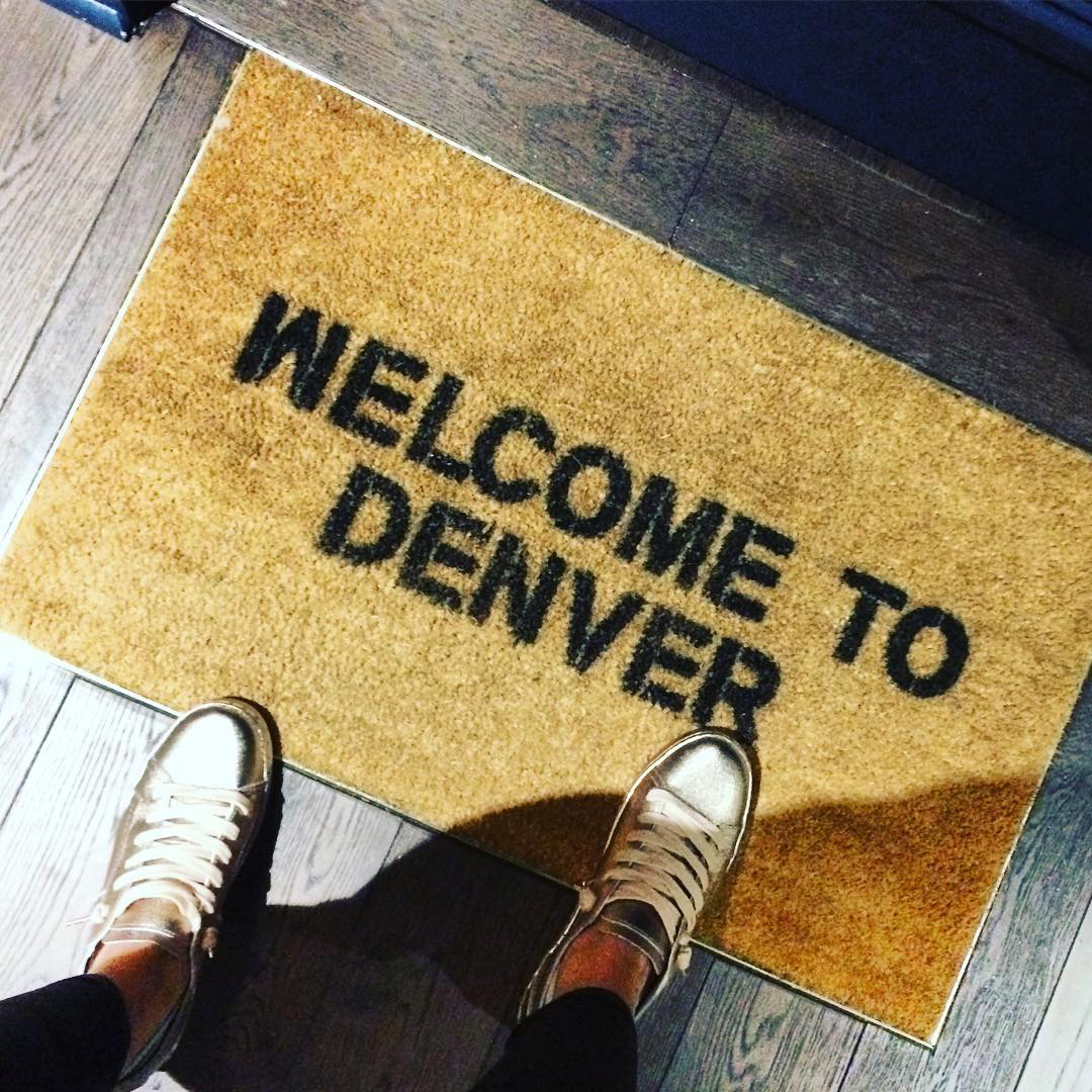 A person in silver sneakers steps onto a tan doormat that says 'Welcome to Denver'