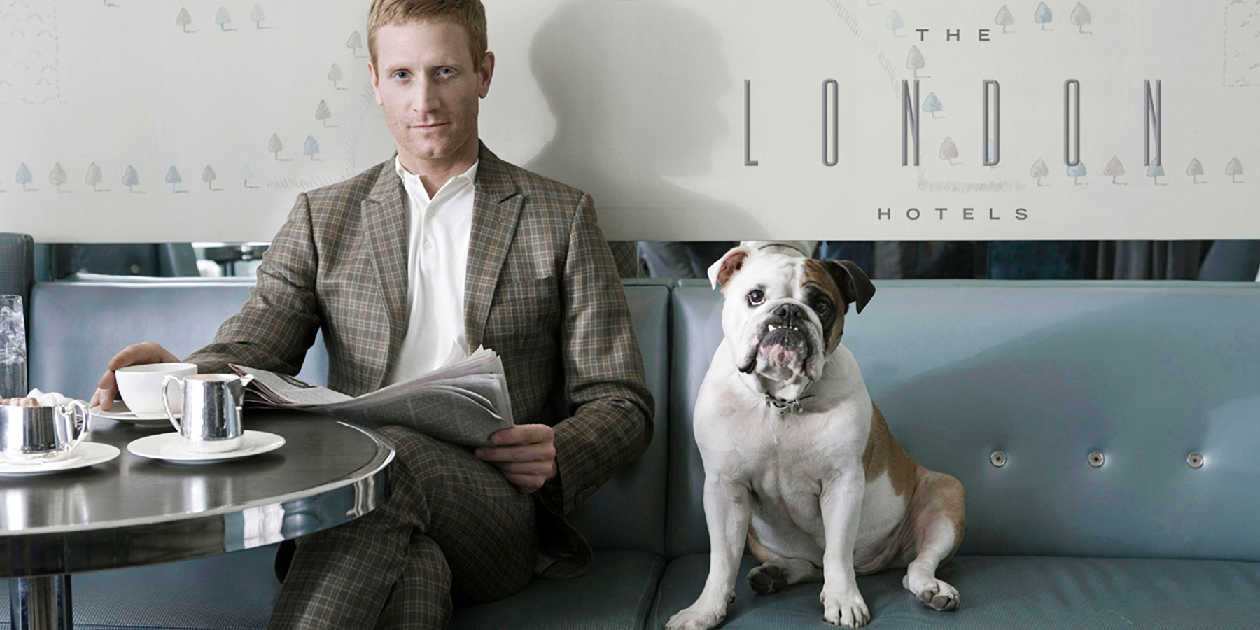 An ad for The London Hotels depicts a man in a suit sitting on a bench seat in a dinner next to a white and brown bulldog