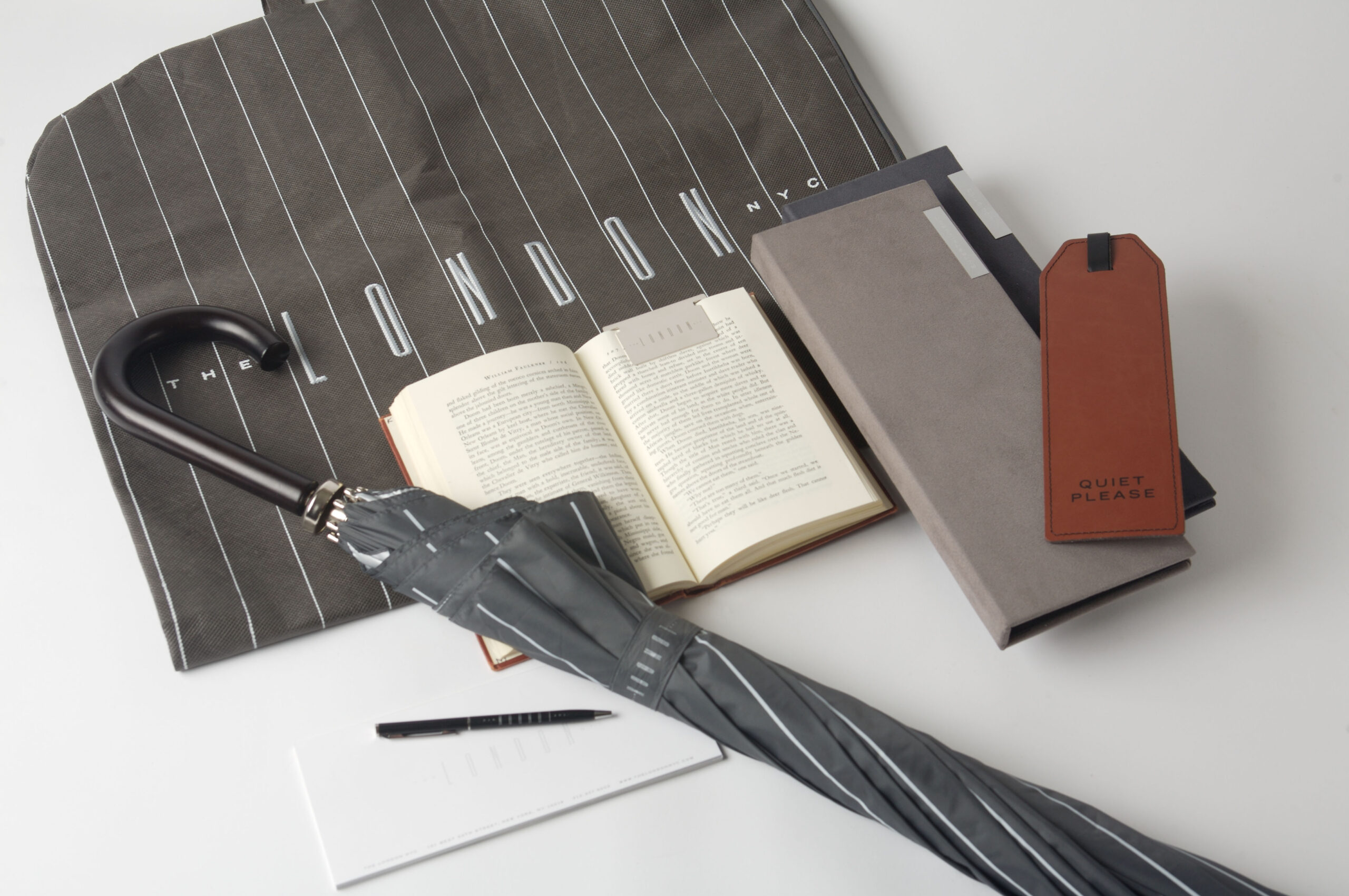 An assortment of items are arranged on a table: an umbrella, a black pinstriped bag embroidered with the words The London NYC, an open book, two narrow binders, a notebook and pen, and a brown leather sign displaying the words Quiet Please