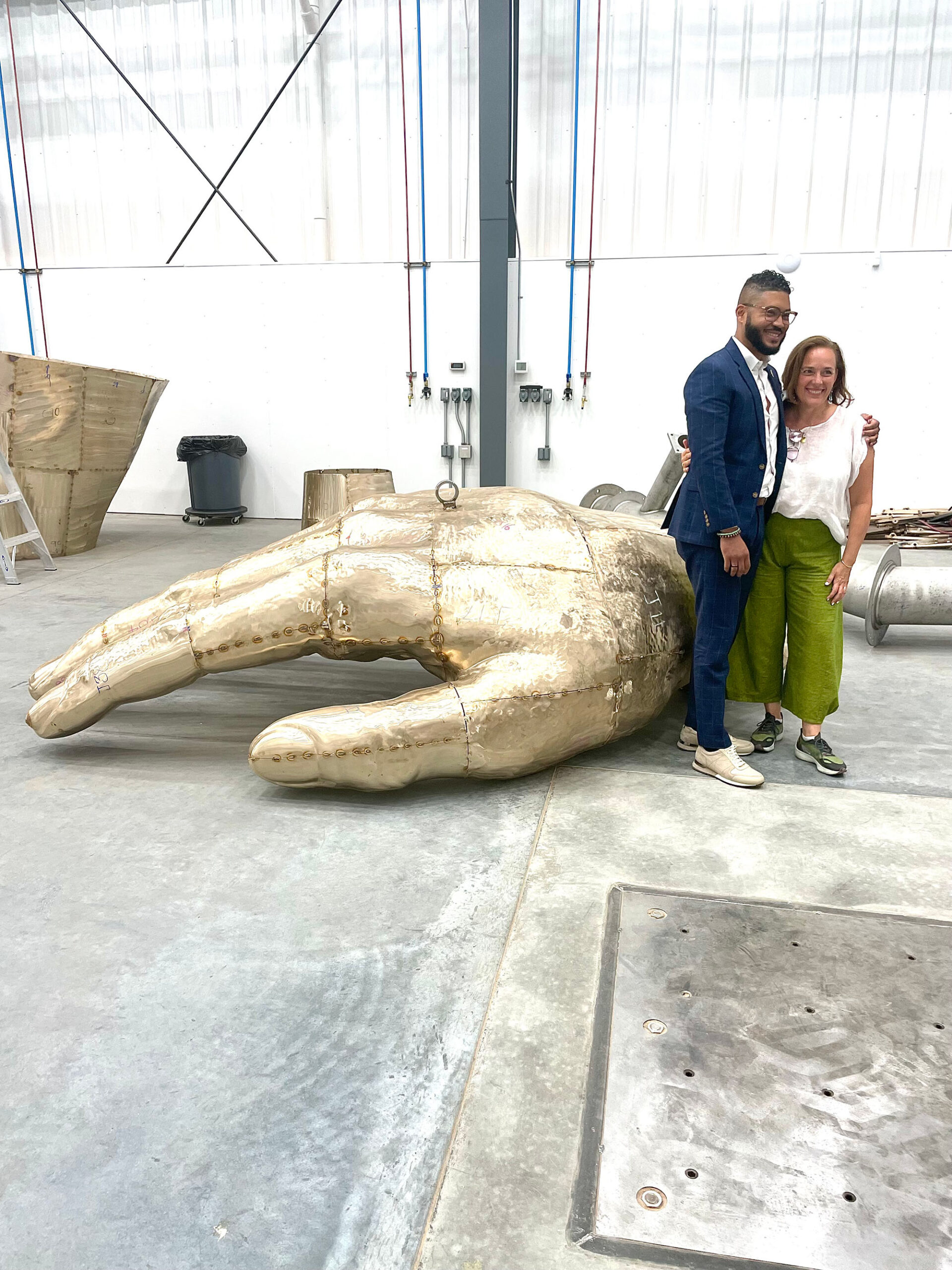 A man and a woman pose for a photo in front of a large golden sculpture of a hand.Embrace Boston, The Embrace Memorial (2023), Imari Paris Jeffries (executive director) and Denise Korn (senior advisor for arts and culture) visit the Walla Walla Foundry during fabrication.