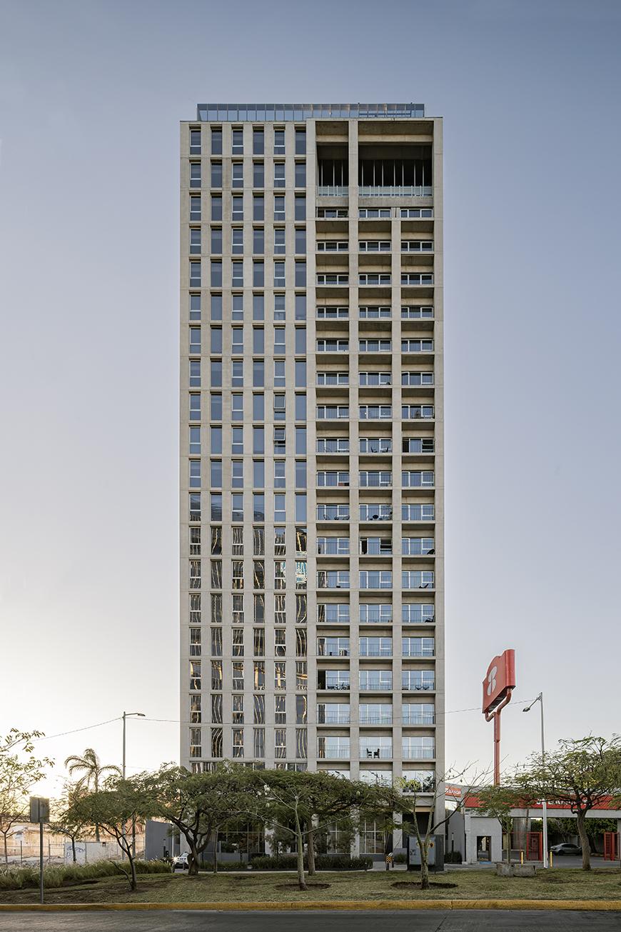 A tall, beige high-rise building with many windows and balconies. The building has a modern design with a flat roof. It is surrounded by trees and a street with parked cars.