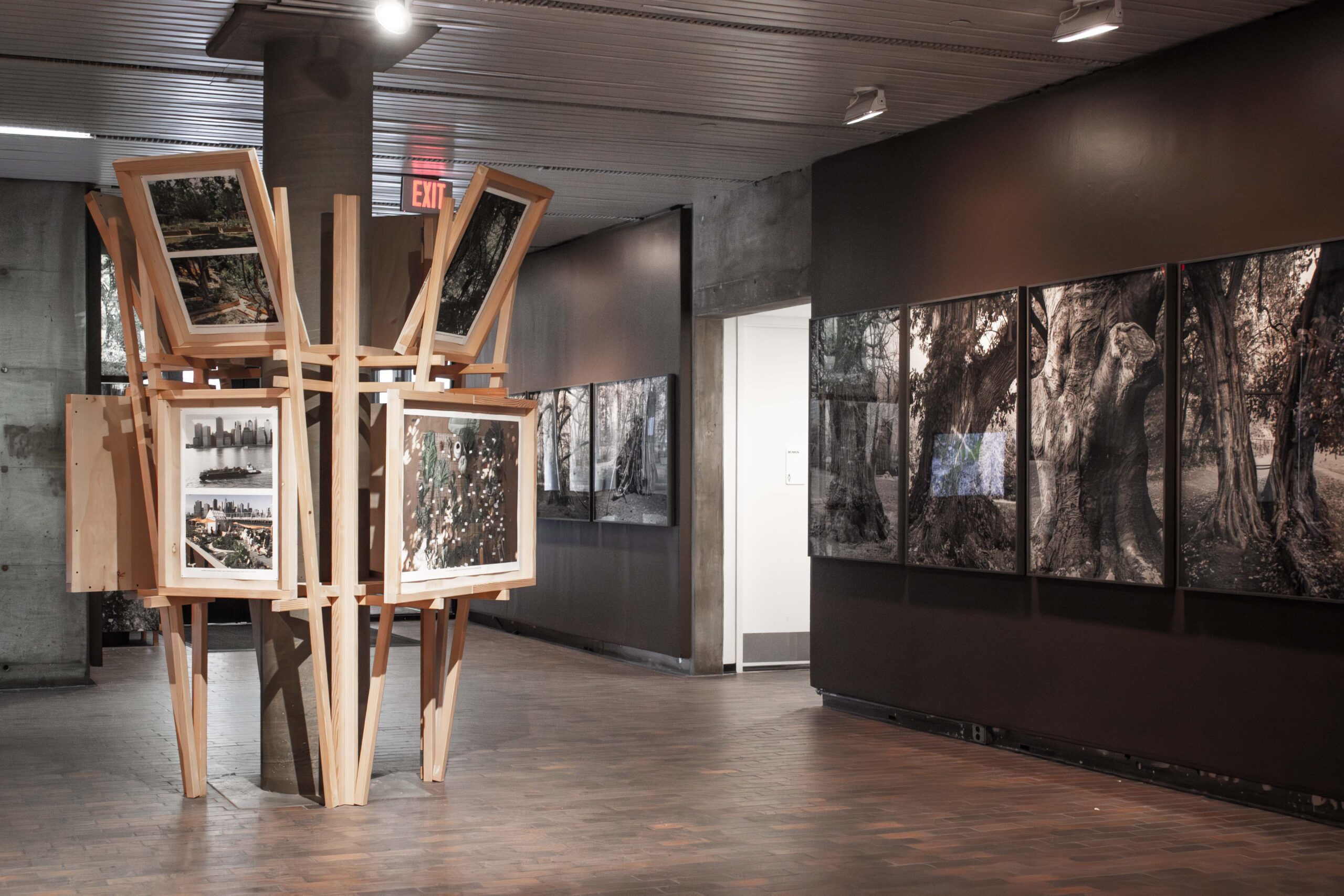 Exhibition featuring gray walls covered with large black and white photographs and a center structure of timber framing with smaller photographs.