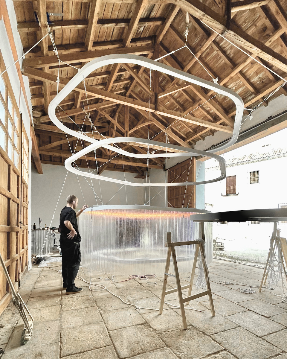 A person stands in a room featuring open wooden beams and a tiled floor with wooden frame sculptures in the center.
