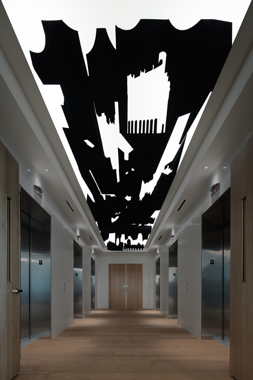 A long, modern hallway with white walls, a brown wooden floor, and a black and white abstract artwork on the ceiling. There are several elevators lined up along one side of the hallway.