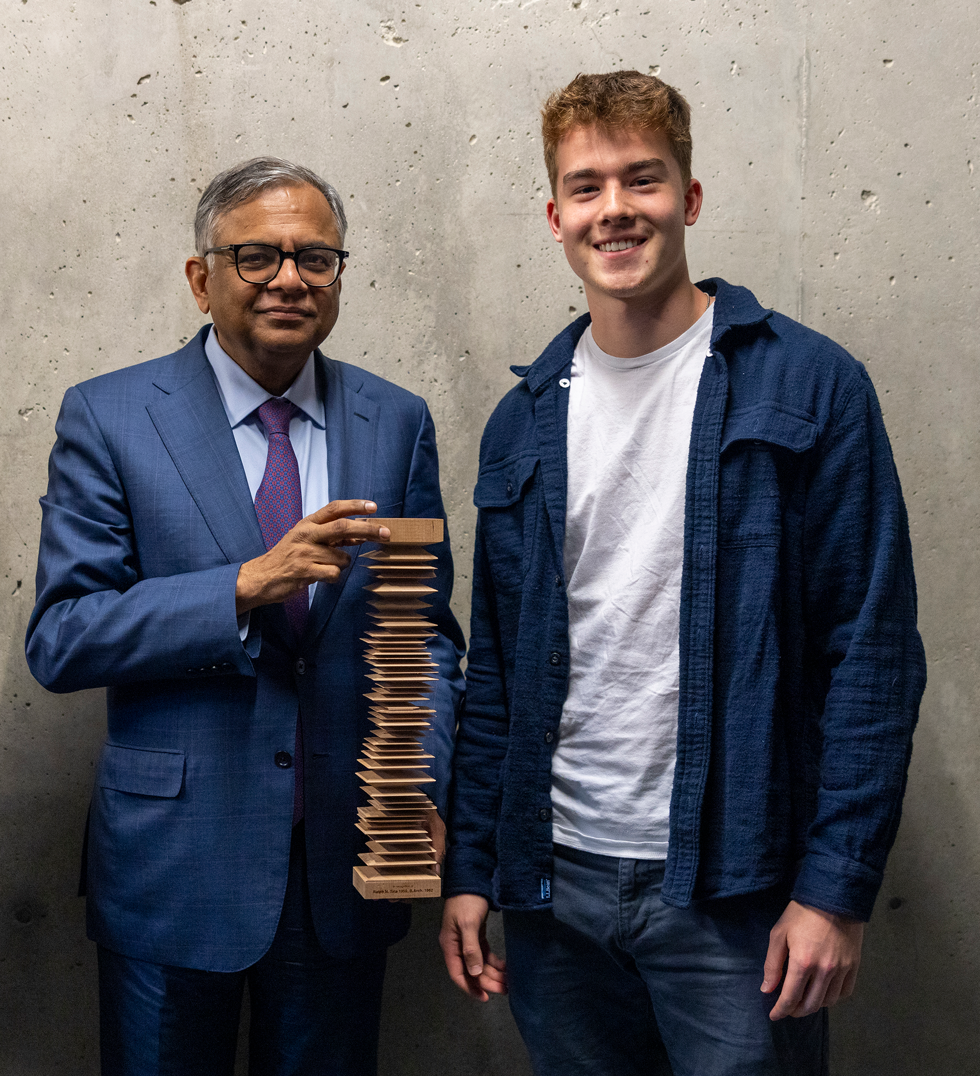 Two smiling people, one holding a wooden award, standing against a concrete background