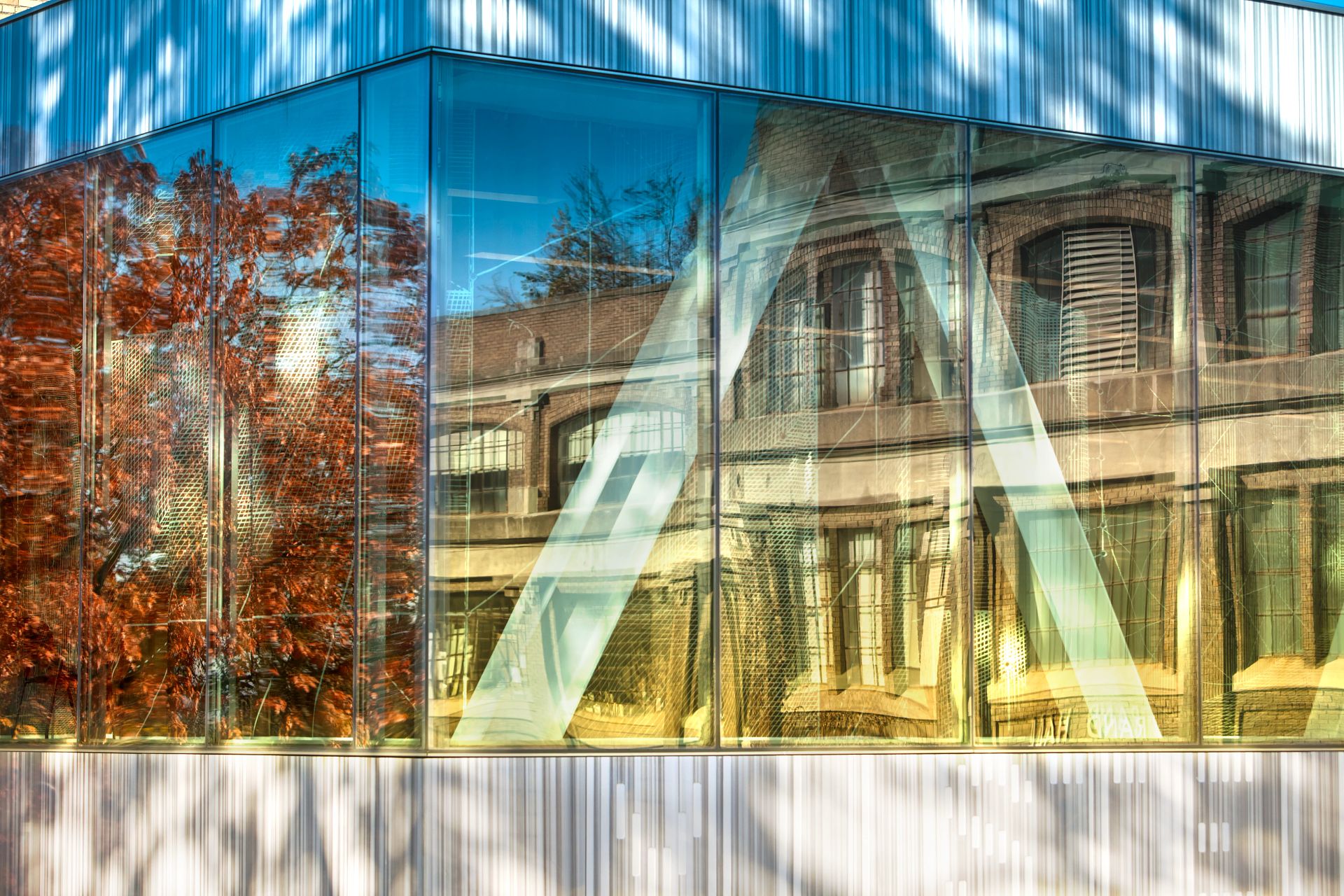 a windowed building corner reflecting the adjacent scenery of fall trees and nearby buildings.