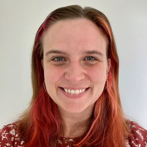 A person smiling with long brown hair with red highlights wearing a red floral top