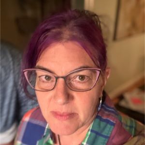 A person with magenta colored hair wearing cat eye glasses standing inside.