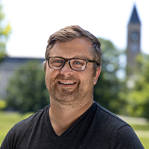 A person with glasses wearing a black v-neck shirt with a clock tower in the distance.