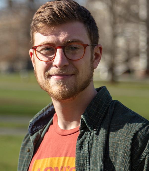 a young man with red hair, red glasses, and a red and yellow t-shirt under a dark green dress shirt