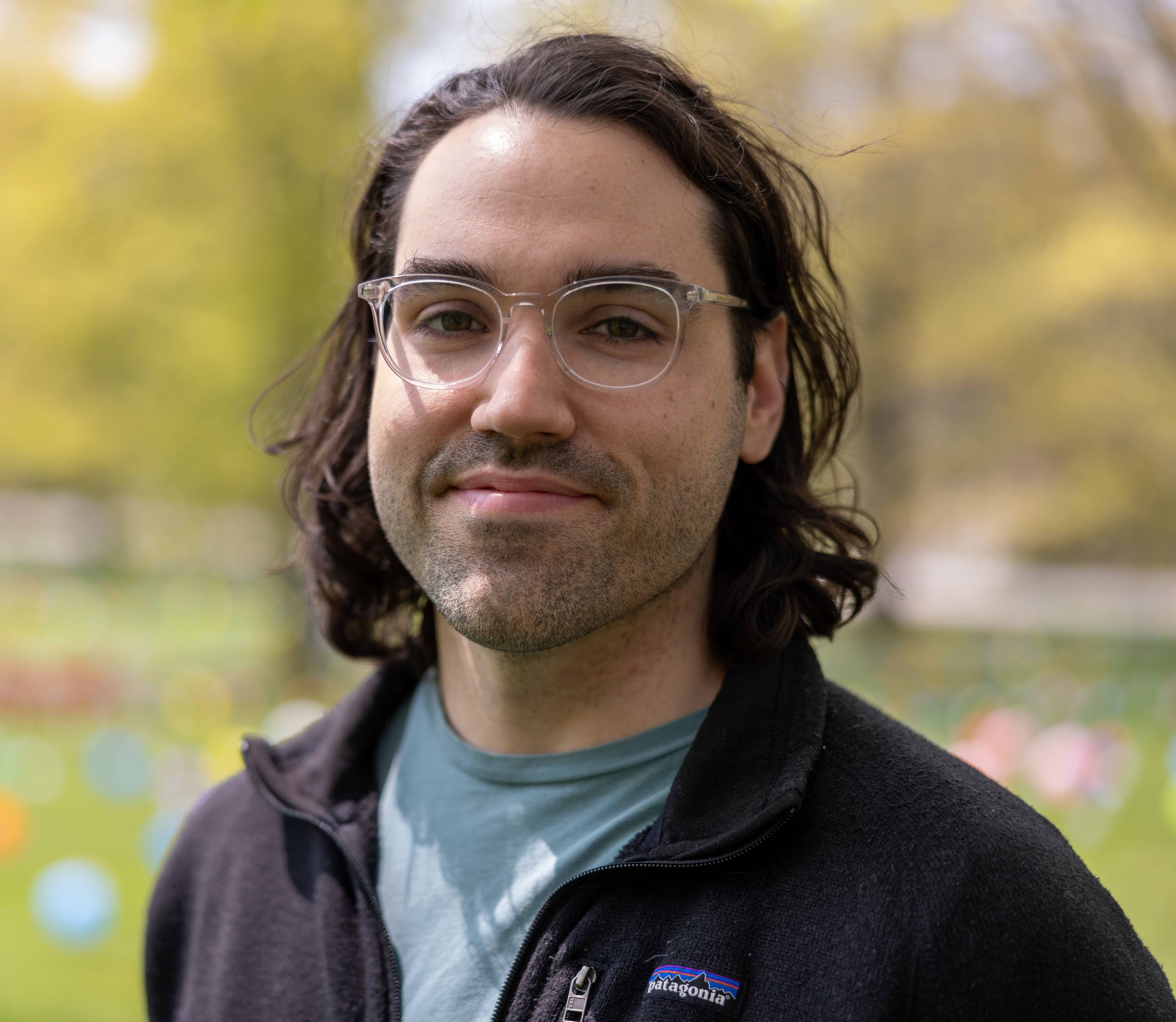 A man with dark long hair wearing clear framed glasses with a t-shirt and jacket