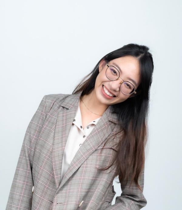 A person with long dark hair, wearing a suit jacket, glasses, smiling and standing in front of a white wall