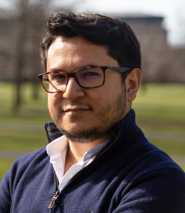 a young man with a beard and brown striped glasses wearing a dark blue quarter zip sweater and a white dress shirt