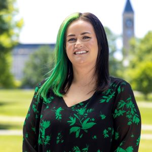 A person with dark hair and green highlights wearing a dark top with green leaves standing outside
