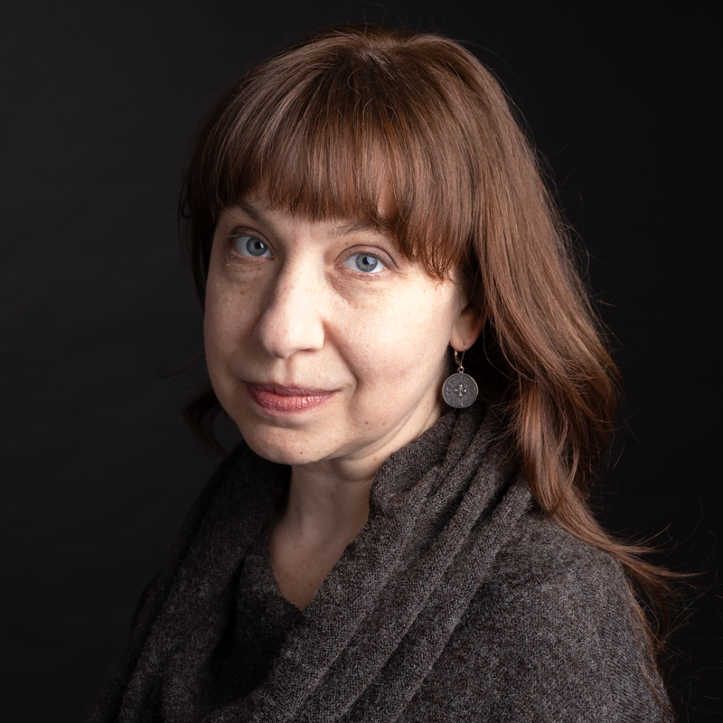 A person with reddish brown hair wearing a brown sweater in front of a black background
