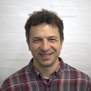 A person smiling with short black hair wearing a plaid button-down shirt standing in front of a white brick wall