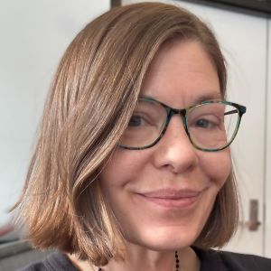 Photo of person with light brown hair cut chin-length. They are smiling at the camera and they are wearing thin black semi-round framed glasses. Their hair is partially obscuring one of their eyes.