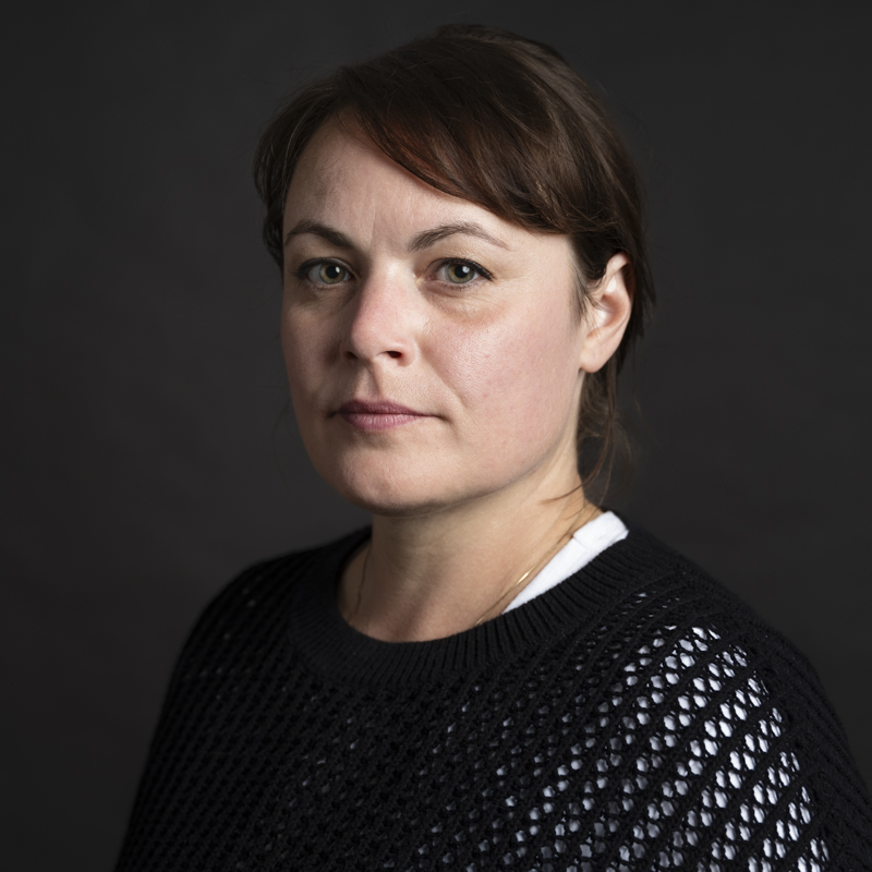 a person with brown hair and a black textured shirt in front of a dark background.